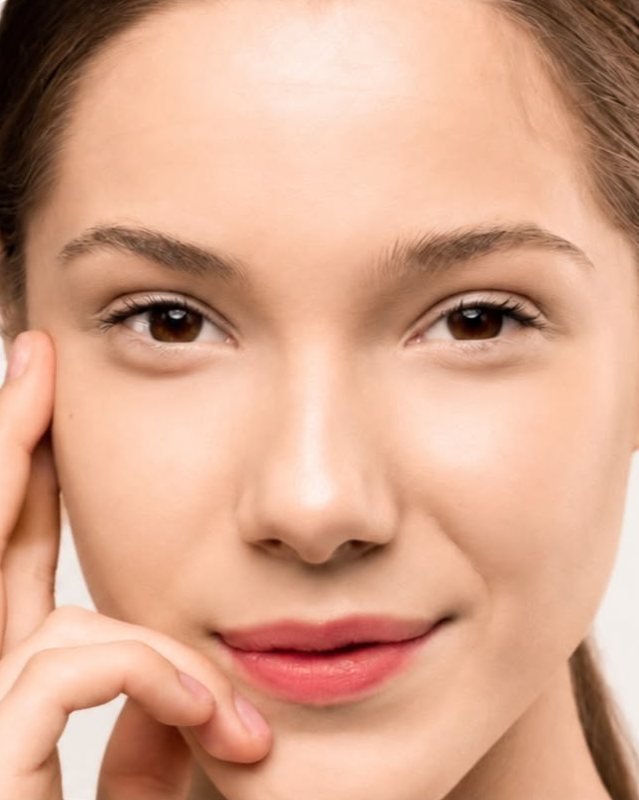 Woman with smooth, glowing skin touching her temple against a soft light backdrop