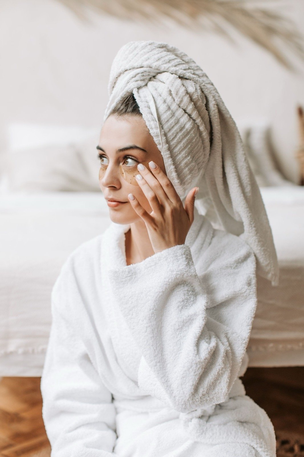 Woman in a white towel wrap and robe gently caring for the skin under her eyes